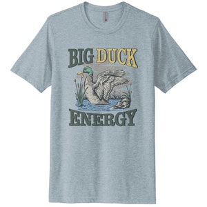 May include: A light blue t-shirt with a vintage-style graphic of a duck in a pond. The text "BIG DUCK ENERGY" is printed in yellow and green letters.