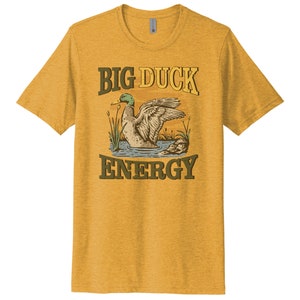 May include: A yellow t-shirt with a vintage-style illustration of a duck on a pond. The text "BIG DUCK ENERGY" is printed in brown letters on the shirt.