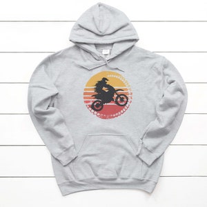 May include: A gray hooded sweatshirt with a vintage-style graphic of a person riding a dirt bike in front of a sunset. The graphic is in shades of orange, red, and yellow.