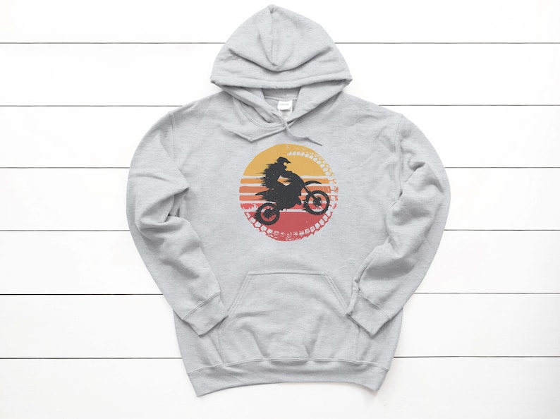 May include: A gray hooded sweatshirt with a vintage-style graphic of a person riding a dirt bike in front of a sunset. The graphic is in shades of orange, red, and yellow.