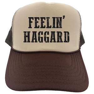 May include: A brown and beige trucker hat with a black mesh back. The front of the hat has the text "FEELIN' HAGGARD" printed in black.
