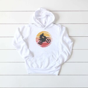 May include: White hooded sweatshirt with a graphic of a person riding a dirt bike in front of a sunset. The graphic is in a circle with a tire tread pattern around the edge.