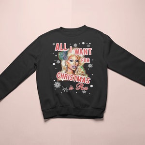 以下が含まれることがあります： 白い雪の結晶のデザインと「All I want for Christmas is RuPaul」というテキストが書かれた黒いスウェットシャツ。