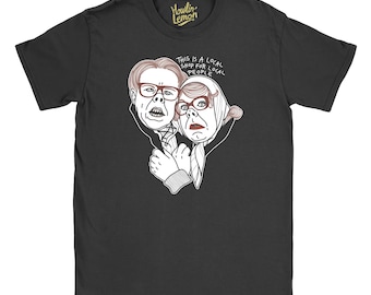 May include: Black t-shirt featuring a cartoon illustration of two people with glasses, one wearing a headscarf. The text "THIS IS A LOCAL SHOP FOR LOCAL PEOPLE" is printed above the illustration. The shirt has a crew neck and short sleeves.