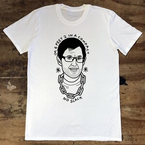 May include: White t-shirt featuring a black and white illustration of a man with glasses and a chain around his neck. The text "I'm 6 feet 2 in a compact * NO SLACK *" is printed on the shirt.