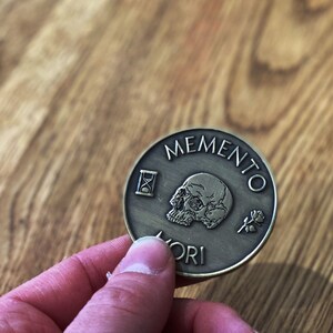 May include: A round, bronze-colored coin with the words "Memento Mori" engraved around the edge. The coin features a skull with a rose and an hourglass.