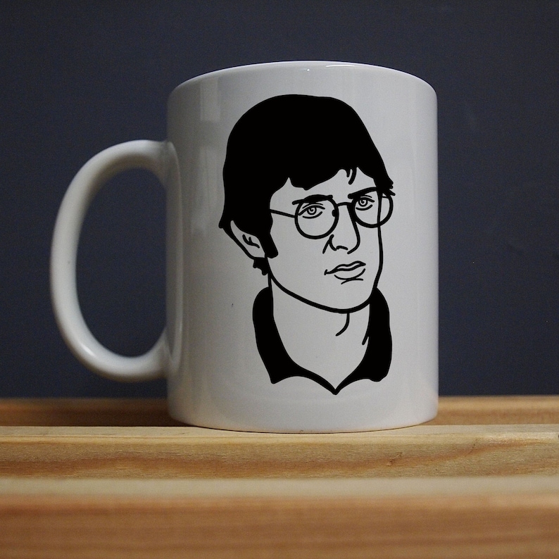 Pu&ograve; includere: Tazza in ceramica bianca con un disegno a tratto nero di una persona con gli occhiali. Il ritratto &egrave; sulla parte anteriore della tazza, con un manico bianco sul lato. La tazza &egrave; su una superficie di legno.