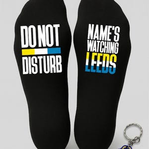 Puede incluir: Calcetines negros con las frases blancas "DO NOT DISTURB" y "NAME'S WATCHING LEEDS". Los calcetines tienen rayas amarillas y azules. También se incluye un llavero con la palabra "ALAW" en azul y amarillo.