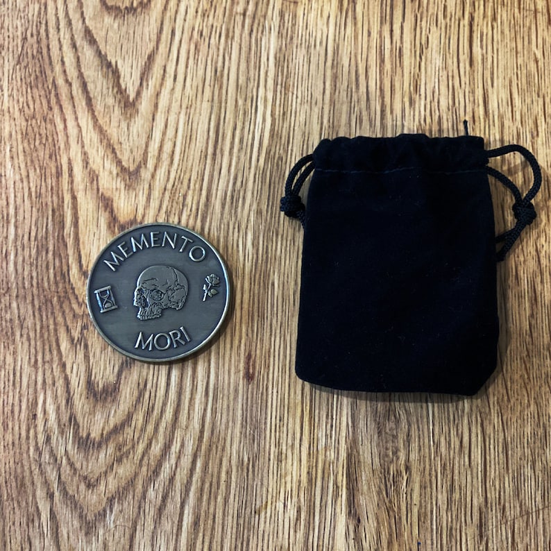 May include: A bronze-colored coin with the words "Memento Mori" and a skull design. The coin is in a black velvet pouch.