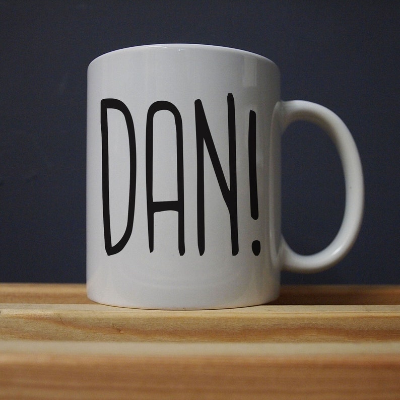 May include: White ceramic mug with a black handle. The mug features the name "DAN!" printed in bold, black capital letters. The mug is sitting on a wooden surface against a dark blue background.