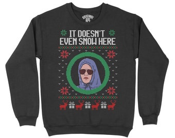 May include: Black sweatshirt with a green and red Christmas sweater design. The design features a person wearing a blue hoodie and sunglasses with the text "It doesn't even snow here".
