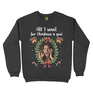 May include: Black sweatshirt with a festive Christmas design. The design includes a woman's face framed by a wreath, red bows, bells, and candy canes. The text "All I want for Christmas is you!" is printed above the wreath.