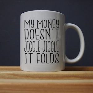 Puede incluir: Taza de cer&aacute;mica blanca con el texto "MY MONEY DOESN'T JIGGLE JIGGLE IT FOLDS" en letras negras. La taza tiene un asa curva y est&aacute; sobre una superficie de madera contra un fondo oscuro. Una idea de regalo humor&iacute;stica.