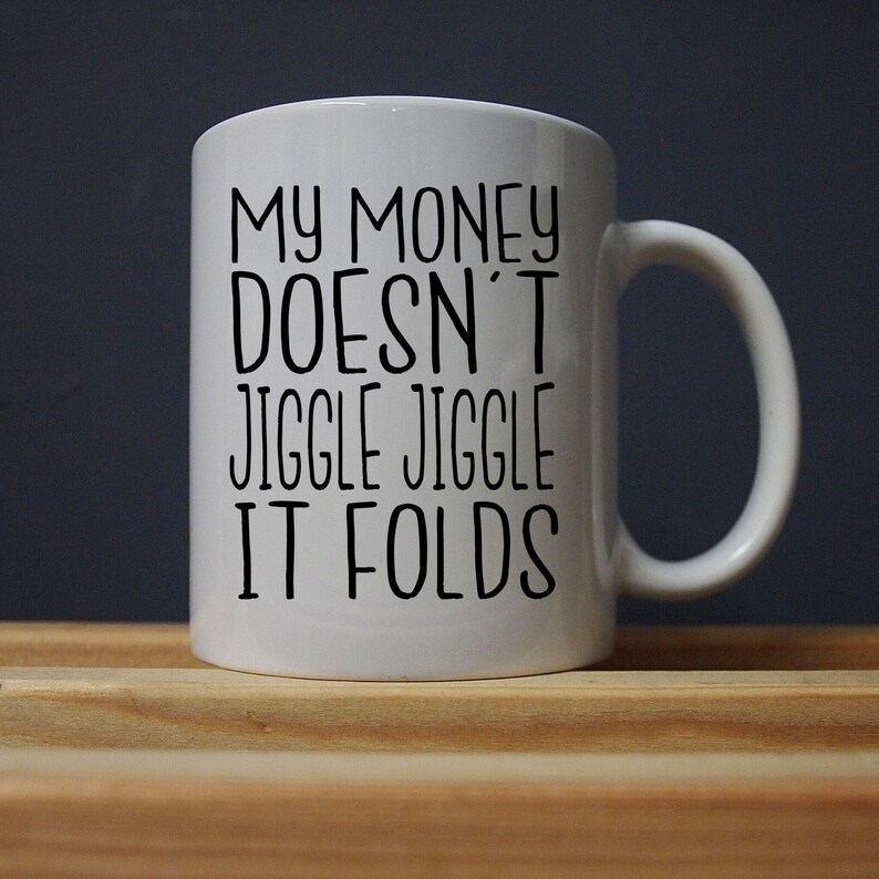 Puede incluir: Taza de cer&aacute;mica blanca con el texto "MY MONEY DOESN'T JIGGLE JIGGLE IT FOLDS" en letras negras. La taza tiene un asa curva y est&aacute; sobre una superficie de madera contra un fondo oscuro. Una idea de regalo humor&iacute;stica.
