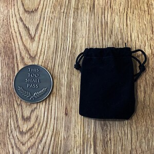 May include: A round, bronze-colored token with the words "This Too Shall Pass" and a laurel wreath design. The token is in a small, black velvet pouch.