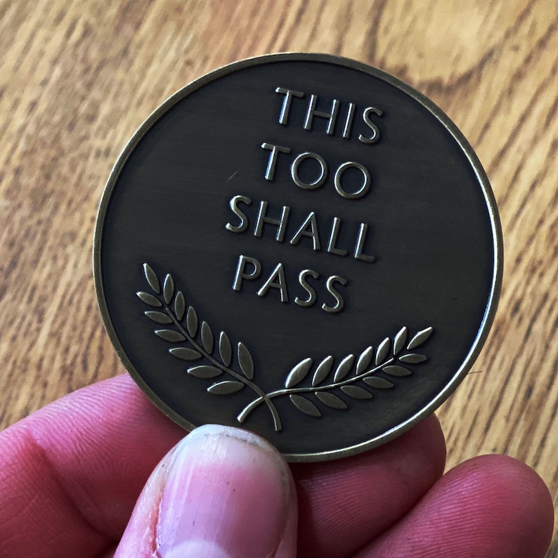 May include: A round, bronze-colored token with the words "This Too Shall Pass" inscribed on it. The words are in a simple, sans-serif font. The token is decorated with a stylized laurel wreath.