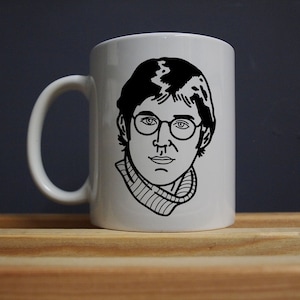 Puede incluir: Taza de cer&aacute;mica blanca con una ilustraci&oacute;n en blanco y negro de una persona con gafas y un cuello alto. La taza tiene un asa curva y una superficie lisa. El dise&ntilde;o es un simple dibujo lineal.