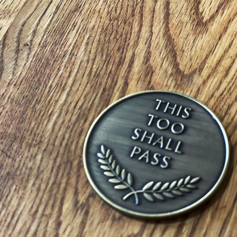 May include: A round, bronze-colored token with the words "This Too Shall Pass" and a laurel wreath design.