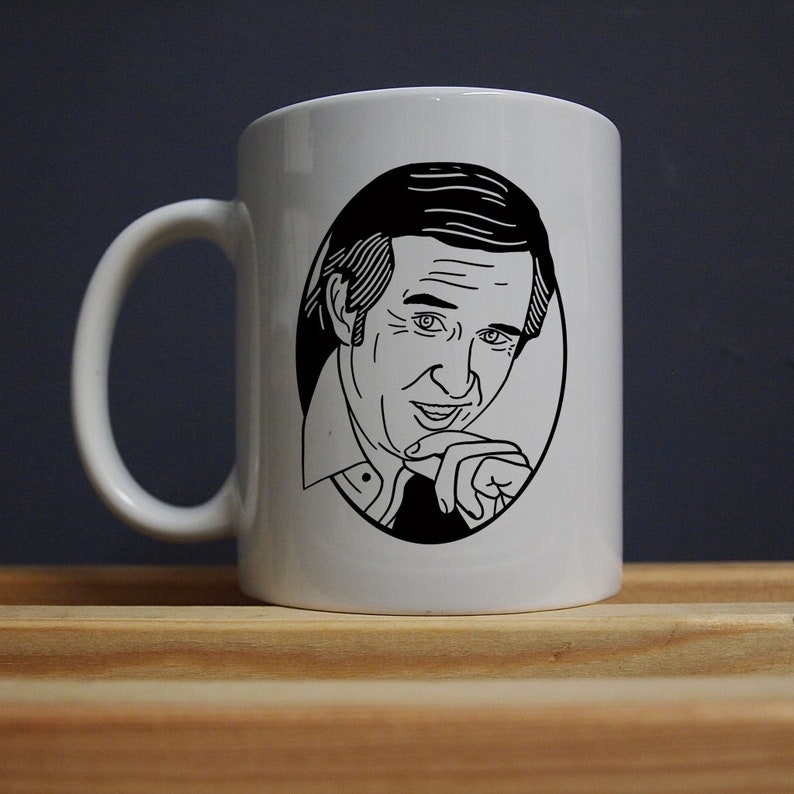 May include: White ceramic mug with a black and white illustration of a man in an oval frame. The man is smiling and touching his chin. The mug has a curved handle and is on a wooden surface.