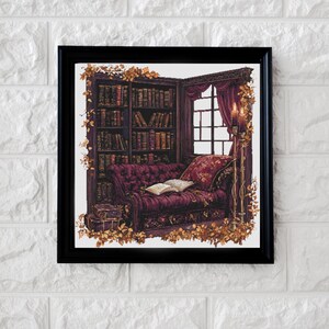 May include: A dark wood framed picture of a cozy reading nook with a burgundy velvet couch, a stack of books, and a lit candle. The couch is decorated with a throw pillow and a book. The room is filled with a warm, inviting atmosphere.