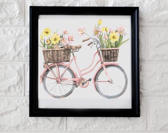May include: A framed watercolor print of a pink bicycle with baskets filled with yellow and pink flowers. The bicycle has a brown seat and handlebars. The artwork is framed in a black frame and set against a white brick wall.