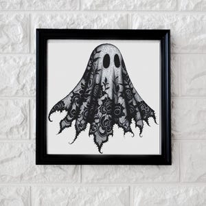 May include: Black framed art print featuring a ghost design. The ghost is depicted with a black lace-like pattern over a white background, with two oval eye shapes. The artwork is mounted on a white brick wall.