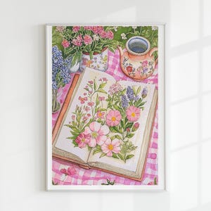 May include: Watercolor painting of an open book with pink flowers and greenery. A teapot, vases of flowers, and lavender sprigs sit on a pink and white checkered tablecloth. The artwork is framed.