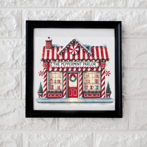 May include: Framed print of "The Peppermint Parlor" shop. The shop has a red and white striped awning and roof, with a red door and window displays. The text "THE PEPPERMINT PARLOR" is above the windows. The frame is black.