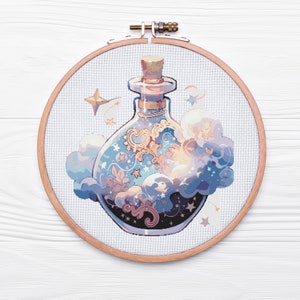 Puede incluir: Un aro de bordado redondo de madera con un diseño de punto de cruz de una botella de vidrio llena de nubes giratorias, estrellas y detalles dorados. La botella tiene un tapón de corcho y está rodeada de un diseño de nubes azul claro y blanco.