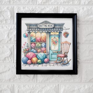 Könnte beinhalten: Ein gerahmter Aquarelldruck eines Strickladens, "Knitting Nook", mit einer blauen Tür, einem Schaukelstuhl und bunten Wollknäueln. Das Schaufenster des Ladens zeigt Wollknäuel in verschiedenen Farben, darunter Rosa, Blau und Gelb.