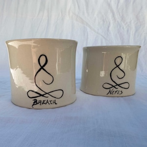 Peut inclure: Deux tasses en céramique blanche avec des dessins au trait noir d'une personne en position de méditation assise. La tasse de gauche dit "Breath" et la tasse de droite dit "Nefes".