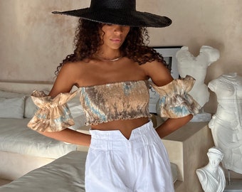 May include: A woman wearing a white off-the-shoulder top with a brown and blue tie-dye pattern, white high-waisted shorts, and a wide-brimmed black hat.