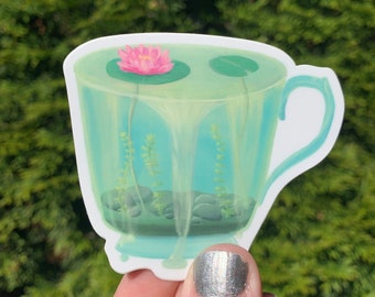 May include: A die-cut sticker featuring a translucent light blue teacup filled with water, containing a pink water lily on a green lily pad, submerged aquatic plants, and small dark gray pebbles resting at the bottom of the cup.