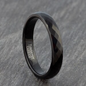 May include: A black tungsten carbide ring with a bevelled edge and the words "TUNGSTEN CARBIDE" engraved on the side.