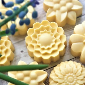 May include: Five handmade soap bars shaped like flowers. The soaps are a pale yellow colour and have intricate floral designs. The soaps are arranged on a light wood surface with a sprig of blue flowers.