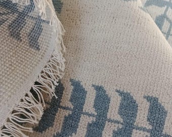 May include: Close-up of a rolled-up rug with a repeating leaf pattern in light blue on a cream background. The rug has a textured surface and a fringed edge. The letters "WOS" are visible on the rug.
