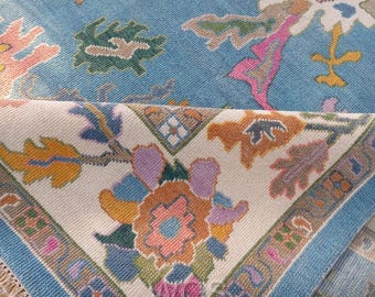 Könnte beinhalten: Nahaufnahme eines blauen Teppichs mit floralem Muster. Der Teppich hat einen hellblauen Hintergrund mit farbenfrohen Blumenmustern in Orange, Rosa, Grün und Lila. Ein dunkelblauer Rand mit floralen Motiven und Fransen umrahmt das Design.