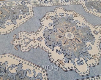Puede incluir: Una alfombra de color azul claro y crema con un intrincado diseño geométrico. La alfombra presenta un diseño de medallón central y bordes decorativos. La alfombra tiene el texto "WOVEN OUSHAK STUDIO" en la esquina superior izquierda y "WOS" en la inferior derecha.