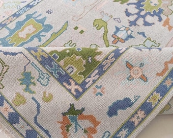 May include: A light blue area rug with a detailed pattern in shades of green, orange, and blue. The rug features a border with geometric designs and the text "WOS" in the corner. The rug is partially rolled up, revealing its underside.