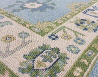 May include: A light blue and cream-colored area rug with a floral pattern. The rug features a central field of light blue with large, stylized floral motifs in shades of blue, green, and cream. The border is cream with green and blue floral designs.