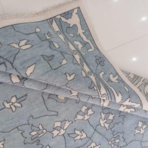 Op de afbeelding: Een lichtblauw tapijt met een bloemenpatroon en een crèmekleurige rand. Het tapijt heeft een herhalend ontwerp van gestileerde witte en grijze bloemen en bladeren. Het tapijt is gedeeltelijk opgevouwen en onthult de textuur en het ontwerp.