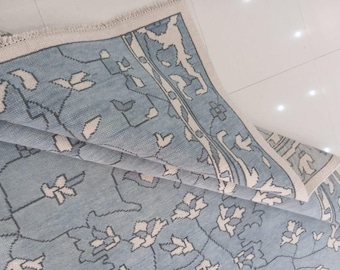 May include: A light blue rug with a floral pattern and a cream-colored border. The rug features a repeating design of stylized flowers and leaves in white and gray. The rug is partially folded, revealing its texture and design.