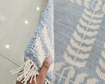 May include: A light blue and cream area rug featuring a repeating geometric pattern with a textured, tufted pile. The rug has a folded corner revealing the underside and white fringe tassels along the edge. A logo reads Woven Oushak Studio.