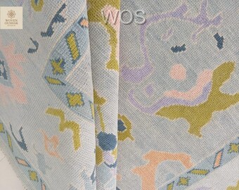 May include: Close-up of a light blue woven rug with a geometric pattern. The design incorporates shapes in pink, yellow, blue, and purple. The rug has a textured appearance and the letters "WOS" are visible.