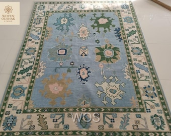 May include: A rectangular, light blue rug with an intricate floral pattern. The rug features a central design of stylized flowers in shades of green, pink, and beige, surrounded by a border with repeating floral and geometric motifs. The rug has the text "WOVEN OUSHAK STUDIO" in the top left and "WOS" in the bottom right.