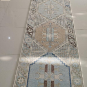 Op de afbeelding: Een rechthoekig vloerkleed met geometrische patronen in lichtblauw, beige en bruin. Het kleed heeft een centraal medaillonpatroon. Dit geweven kleed is geschikt voor gangen of entrees. De lengte is ongeveer 200 cm.