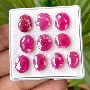 Pode incluir: Uma coleção de nove gemas de rubi facetadas de forma oval em uma caixa branca. Os rubis são de cor rosa intenso e têm um alto nível de brilho.