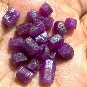 May include: A collection of rough, deep purple ruby gemstones. The unpolished stones have a natural, crystalline texture. The gems vary in size and shape, with facets catching the light. These rubies are a rich, vibrant colour.