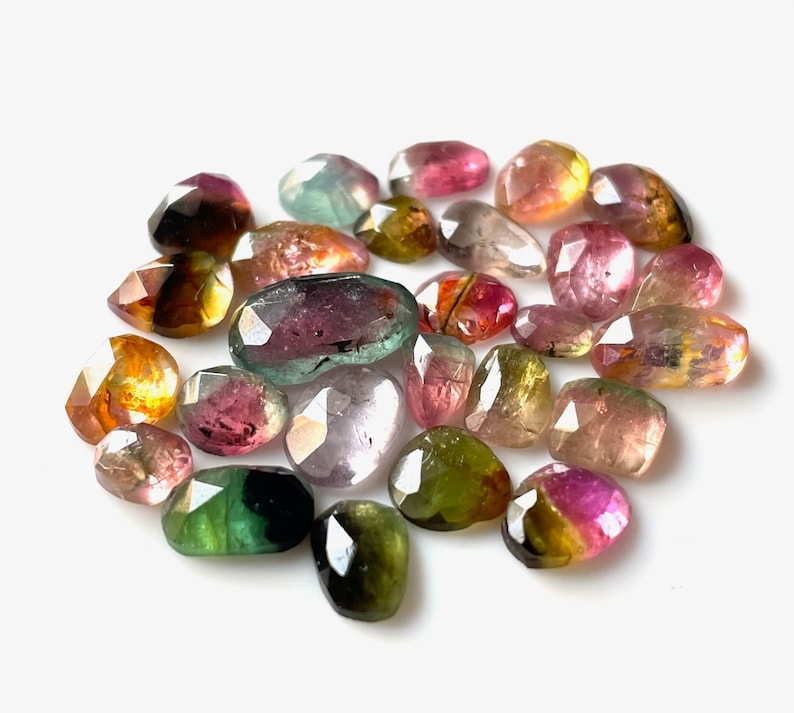 May include: A collection of 20 faceted, multi-colored gemstones. The stones are various shades of green, pink, yellow, and brown. The stones are arranged in a loose circle on a white background.