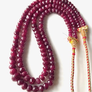 Könnte beinhalten: Eine mehrsträngige Halskette aus facettierten, tiefroten Edelsteinen. Die Halskette hat eine geflochtene Kordel mit goldenen und roten Akzenten an den Enden. Die Edelsteine haben eine polierte, reflektierende Oberfläche, vor weißem Hintergrund.